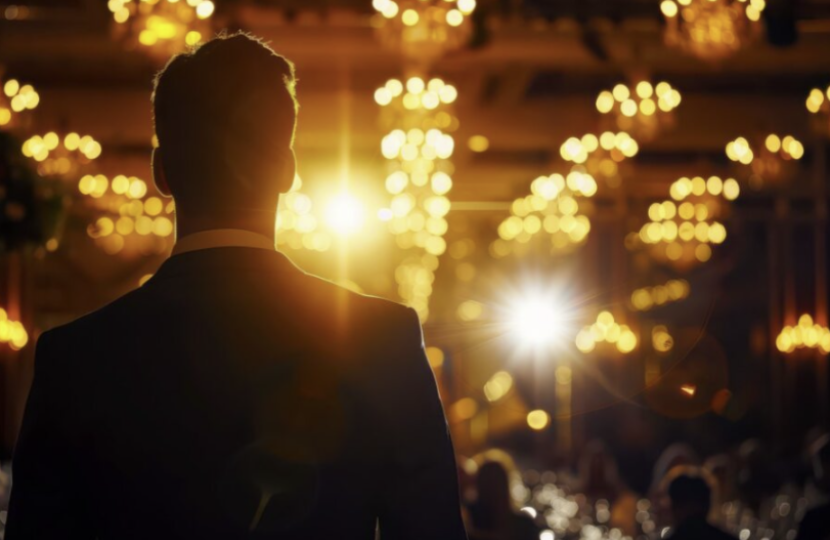 A man in a suit stands with his back to the camera, facing a brightly lit, elegant ballroom in London Mayfair, filled with chandeliers and blurred lights, suggesting he is about to address an audience at a political event.